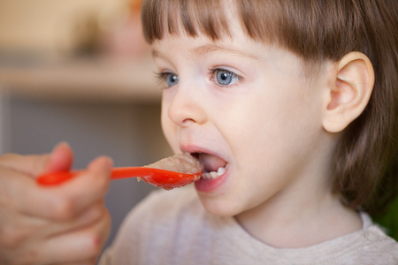 Beautiful baby eats porridge from moms hand. The mother feeds the son with a spoon. Boy with long blond hair and big blue eyes probablyの写真素材
