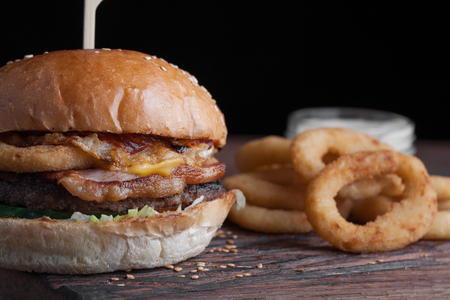 Closeup of a tasty Burger with appetizers such as fried onion rings with a white garlic sauce. Juicy Burger with bacon and cheese on a dark wood background.の写真素材