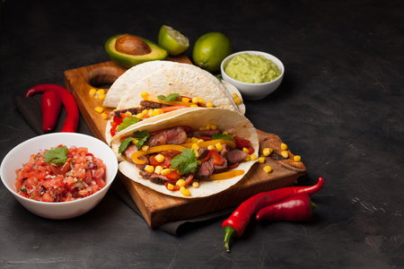 Three Mexican tacos with marbled beef, black Angus and vegetables on wooden Board on a dark stone background. Mexican dish with sauces guacamole and salsa in bowls. top view.の写真素材