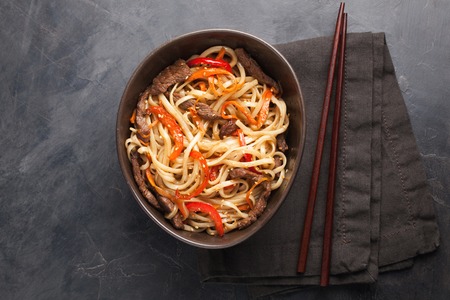 Close-up of noodles in a glass bowl with vegetables and beef in teriyaki sauce on a dark concrete background. Top view of Asian food, and wooden sticks. The concept of fast food.の写真素材