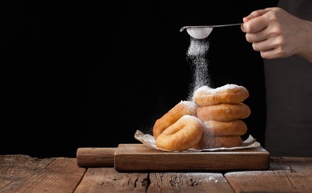 Baker sprinkles sweet donuts with powder sugar on black background. Delicious, but unhealthy food on the old wooden table with copy space.の写真素材