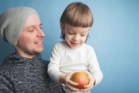 Dad plays with a lovely little son, sitting in his arms on a blue background in the studio. It...