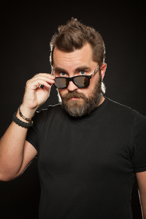 A handsome, strong man with a stylish hairstyle and beard dresses sunglasses in the Studio on a black background.の写真素材