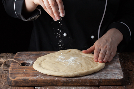 Chef hands pouring flour powder on raw dough using sieve on a black background, Cooking process.の写真素材