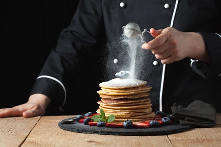 Serving pancakes with powdered sugar and berries. Chef woman hand. Beautiful food still life. slightly toned image, dark black background with text area. Horizontal view.の写真素材