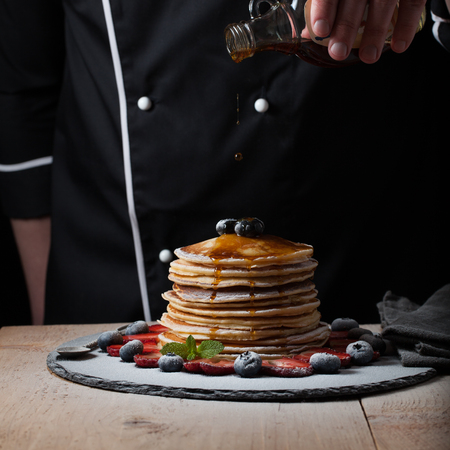 The chef pours maple syrup on pancake stack with blueberries and strawberries on black background.の写真素材