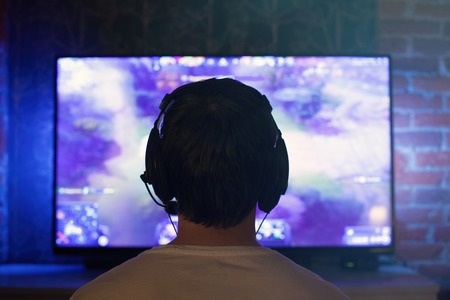 Gamer or streamer in earphones with microphone sits at home in dark room and plays with friends on networks in video games. A young man sits in front of a monitor or TV.の写真素材