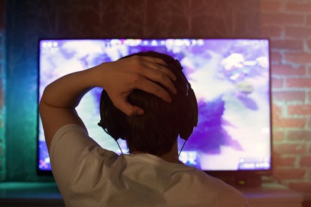 Gamer or streamer in earphones with microphone sits at home in dark room and plays with friends on networks in video games. The young man is angry and grabs his head, as his team lost the game.の写真素材