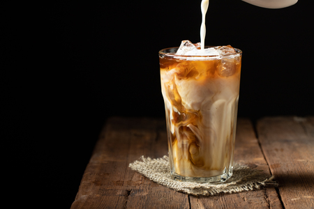 Ice coffee in a tall glass with cream poured over and coffee beans on a old rustic wooden table. Cold summer drink on a dark wooden background with copy space.の写真素材