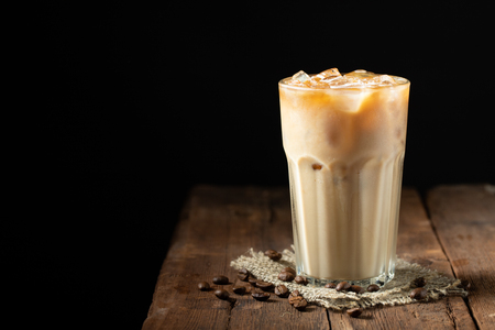 Ice coffee in a tall glass with cream poured over and coffee beans on a old rustic wooden table. Cold summer drink on a dark wooden background with copy spaceの写真素材