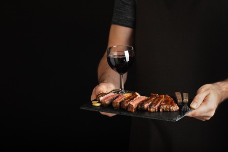 Man holding juicy grilled beef steak with spices and red wine glass on a stone cutting board on a black background. With copy space for text.の写真素材