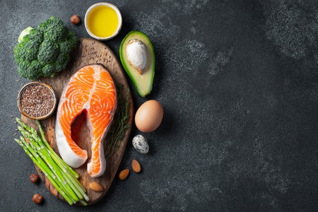 A set of healthy food for keto diet on a dark background. Fresh raw salmon steak with flax seeds, broccoli, avocado, chicken eggs, nuts and asparagus on a wooden Board. Top view with copy space.の写真素材