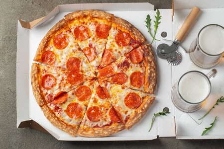 Italian fast food. Delicious hot pepperoni pizza in a box, and glass of beer sliced and served on brown table, close up view. Concept of fast food and unhealthy diet. Top view. Flat lay.の写真素材