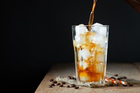 Ice coffee in a tall glass with cream poured over, ice cubes and beans on a old rustic wooden table. Cold summer drink with tubes on a black background with copy spaceの写真素材