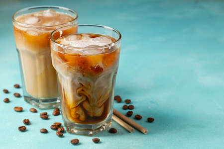 Ice coffee in a tall glass with cream poured over and coffee beans on a light stone background. Cold...