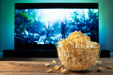 A glass bowl of popcorn and remote control in the background the TV works. Evening cozy watching a...