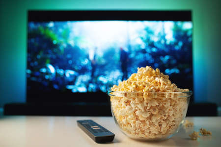 A glass bowl of popcorn and remote control in the background the TV works. Evening cozy watching a movie or TV series at home.の写真素材
