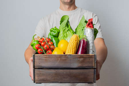 Delivery Concept -Young Caucasian delivery man carrying wooden box of grocery vegetables and fruits from store. Isolated on Gray studio Background. Copy Space.の写真素材