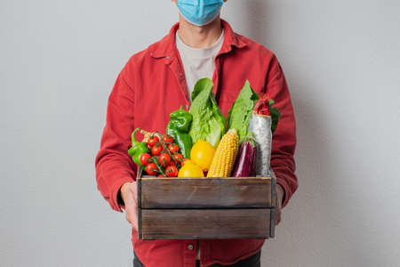 Young delivery man in protective face mask holding wooden box in hands. Man courier with shopping box. Safe online delivery from supermarket to home.の写真素材