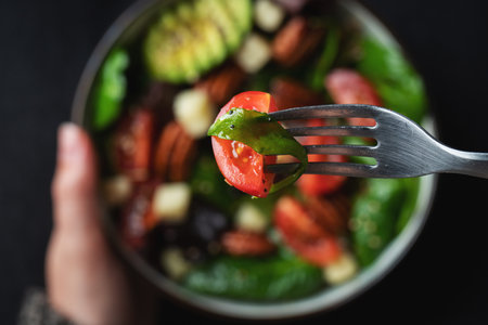 Girl eating healthy homemade salad with tomatoes, cheese cubes, pecans, avocado, spinach and arugula. Green vegetarian meal in a bowl in the girl's hands on a dark background. The concept of healthy eating.の写真素材