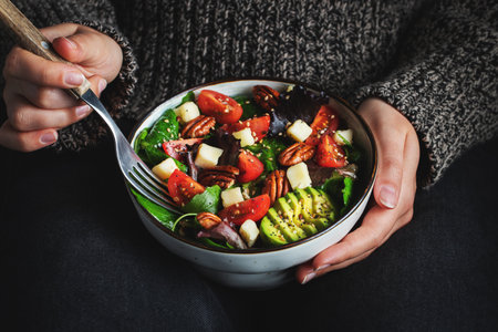 Healthy homemade salad with tomatoes, cheese cubes, pecans, avocado, spinach and arugula. Green vegetarian meal in a bowl in the girl's hands on a dark background. The concept of healthy eating.の写真素材