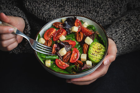 Healthy homemade salad with tomatoes, cheese cubes, pecans, avocado, spinach and arugula. Green vegetarian meal in a bowl in the girl's hands on a dark background. The concept of healthy eating.の写真素材