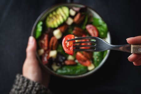 Girl eating healthy homemade salad with tomatoes, cheese cubes, pecans, avocado, spinach and arugula. Green vegetarian meal in a bowl in the girl's hands on a dark background. The concept of healthy eating.の写真素材