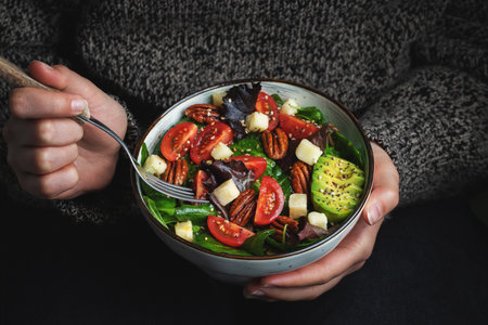 Healthy homemade salad with tomatoes, cheese cubes, pecans, avocado, spinach and arugula. Green vegetarian meal in a bowl in the girl's hands on a dark background. The concept of healthy eating.の写真素材