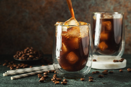 Ice coffee in a tall glass with cream poured over, ice cubes and beans on a dark concrete table. Cold summer drink with tubes on a black background with copy space.の写真素材