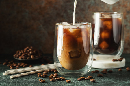 Ice coffee in a tall glass with cream poured over, ice cubes and beans on a dark concrete table. Cold summer drink with tubes on a black background with copy space.の写真素材
