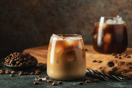 Ice coffee in a tall glass with cream poured over, ice cubes and beans on a dark concrete table. Cold summer drink with tubes on a black background with copy space.の写真素材