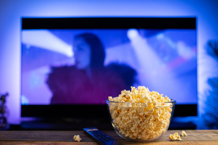 A glass bowl of popcorn and remote control in the background the TV works. Evening cozy watching a...