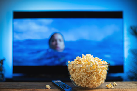 A glass bowl of popcorn and remote control in the background the TV works. Evening cozy watching a...