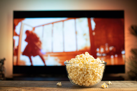 A glass bowl of popcorn and remote control in the background the TV works. Evening cozy watching a...