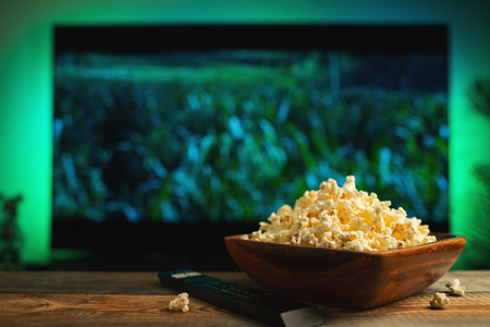 A wooden bowl of popcorn and remote control in the background the TV works. Evening cozy watching a...