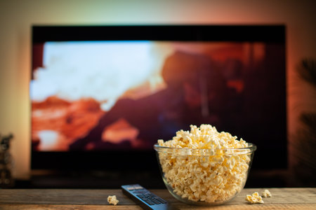 A glass bowl of popcorn and remote control in the background the TV works. Evening cozy watching a movie or TV series at home.の写真素材