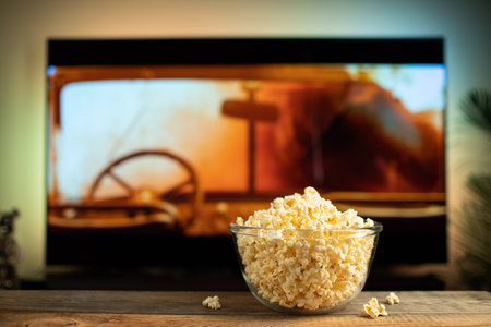 A glass bowl of popcorn and remote control in the background the TV works. Evening cozy watching a movie or TV series at home.の写真素材