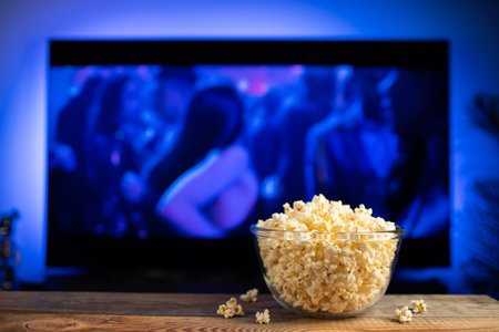 A glass bowl of popcorn and remote control in the background the TV works. Evening cozy watching a...