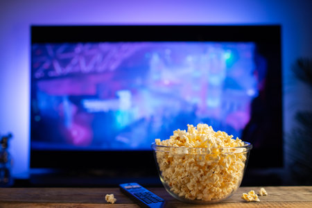 A glass bowl of popcorn and remote control in the background the TV works. Evening cozy watching a...
