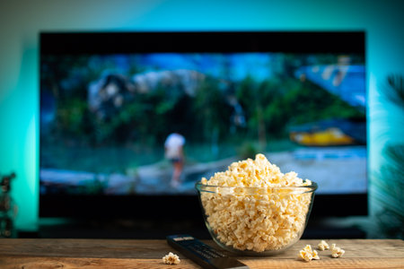 A glass bowl of popcorn and remote control in the background the TV works. Evening cozy watching a movie or TV series at home.の写真素材