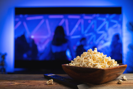 A wooden bowl of popcorn and remote control in the background the TV works. Evening cozy watching a...