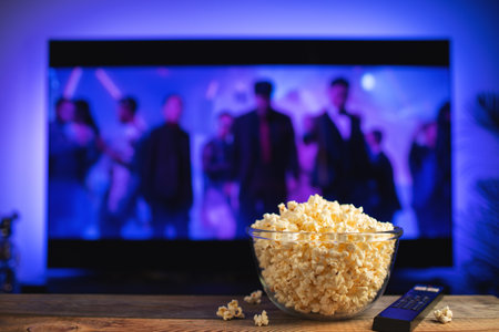 A glass bowl of popcorn and remote control in the background the TV works. Evening cozy watching a movie or TV series at home.の写真素材