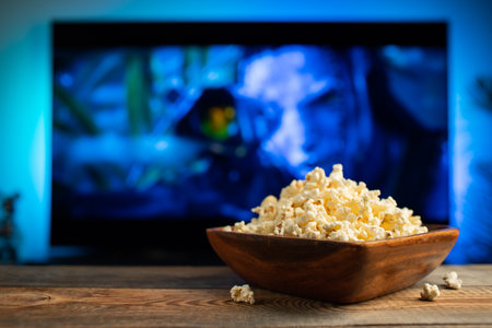 A wooden bowl of popcorn and remote control in the background the TV works. Evening cozy watching a...