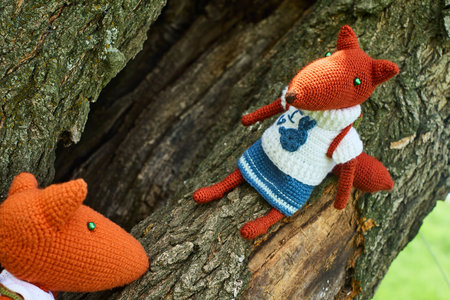 Two knitted fox toys, dressed in clothes and with backpacks, are attached to the bark of a tree. Children's toys, handmadeの写真素材