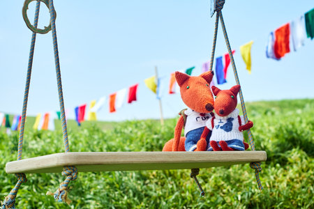 Two crochet fox toys, dressed in clothes and with backpacks, riding on a swing. Children's toys, handmadeの写真素材