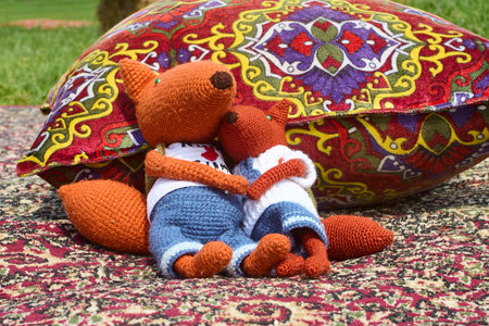 Two soft knitted fox toys, dressed in clothes and with backpacks, snuggle up to each other on a colorful oriental rug next to a throw pillow, creating a cozy atmosphere of warmth aの写真素材