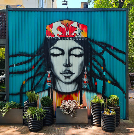 Large vibrant graffiti of a pensive girl in traditional Uzbek headdress and earrings on a blue metal wall. Located in Tashkent, showcasing modern street art with national flair.の写真素材
