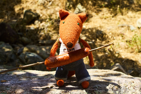 A knitted toy fox holding a roasted sausage on a stick, with a wilderness background. Photographed in July 2025.の写真素材