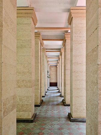 A vertical, symmetrical shot of a long covered walkway, formed by rows of light-colored columns and featuring decorative red and gray tiling on the floor.の写真素材