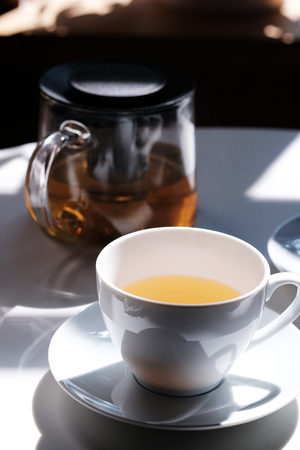 White cup of tea with a tea kettle on the white tableの写真素材